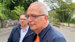 Zwei Männer in blauen Jacken stehen auf einer Straße, im Hintergrund das Schwebebahngerüst, links und rechts grüne Bäume