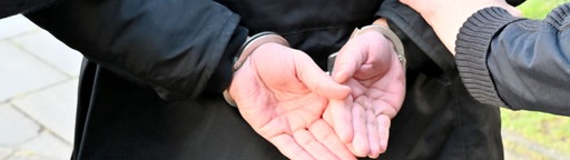 Zwei Männer halten einen Mann fest. Seine Hände sind hinter seinem Rücken in Handschellen.