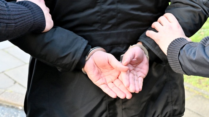 Symbolbild: Zwei Männer halten einen Mann fest. Seine Hände sind hinter seinem Rücken in Handschellen.