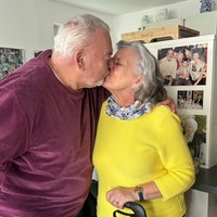 Ein älteres Paar gibt sie einen Kuss. Der Mann hat seinen Arm um die Frau gelegt. Im Hintergrund sind viele Bilder zu sehen.
