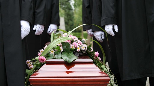 Ein brauner Sagt mit Pink-weißen Blumen drauf, daneben stehen Männer in schwarzen Anzügen und weißen Handschuhen
