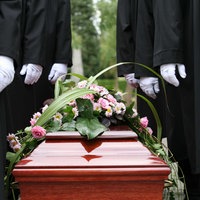 Ein brauner Sagt mit Pink-weißen Blumen drauf, daneben stehen Männer in schwarzen Anzügen und weißen Handschuhen