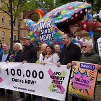Die Initiatoren der Unterschriftenaktion halten ein Banner in den Händen: 100.000 Unterschriften. Im Hintergrund eine Jacques Tilly Figur. Ein großer bunter Fisch mit der Aufschrift "wir sind mehr" frisst einen kleineren AfD Fisch.