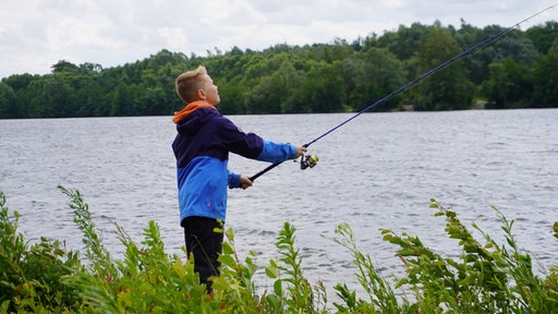Ein Junge steht mit dem Rücken zum Bild und wirft eine Angel aus. Im Hintergrund der Paderbornder Lippesee. Im Vordergrund: Gras und Uferpflanzen.