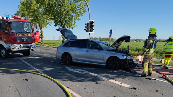 Ein Auto mit zerstörtem Motorraum steht auf der Straße. Daneben sind Einsatzkräfte und ein Feuerwehrwagen.