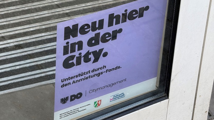 An einer Fensterscheibe hängt ein weißes Schild der Stadt Dortmund mit der Aufschrift "Neu hier in der City? Unterstützt durch den Anmietungs-Fonds."