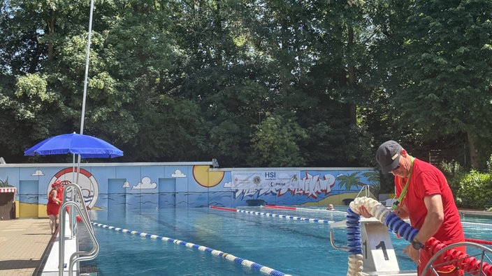 Ein Mann in rotem Shirt und roter Hose steht vor einem Schwimmbecken in einem Freibad. Er unterteilt das Becken in mehrere Bahnen.