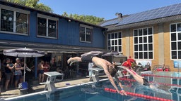 Zwei Menschen springen gleichzeitig mit dem Kopf voran in ein Schwimmbecken. Im Hintergrund sind weitere Menschen zu sehen