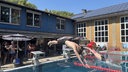 Zwei Menschen springen gleichzeitig mit dem Kopf voran in ein Schwimmbecken. Im Hintergrund sind weitere Menschen zu sehen