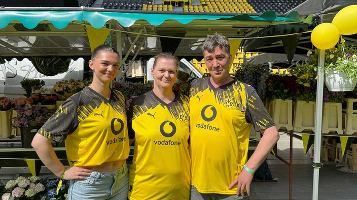 Eine junge Frau neben einer Frau und einem Mann, alle tragen ein BVB-Trikot. Im Hintergrund ein Marktstand.