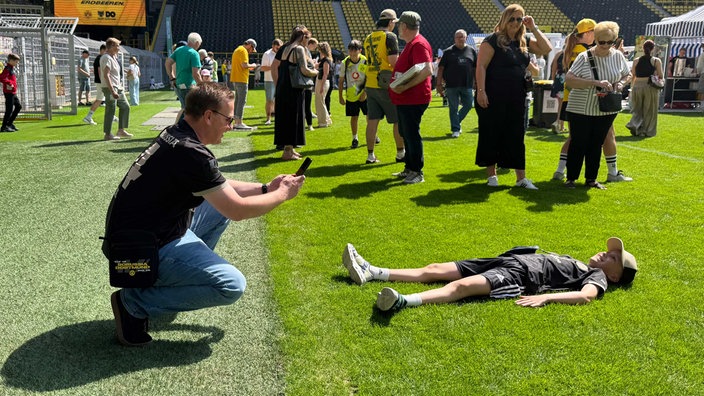 Ein Mann macht ein Foto von einem Jungen, der auf dem Rasen des BVB-Stadions liegt und sich freut.