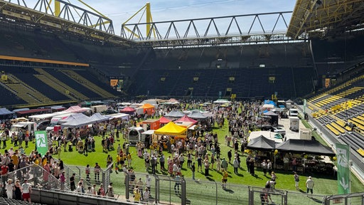 Ein Fußballfeld mit etlichen Marktständen und Besuchern, drum herum leere schwarz-gelbe Tribünen.