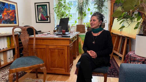 Nahid Farshi sitzt in einer Wohnung, im Hintergrund ein Schreibtisch und Pflanzen