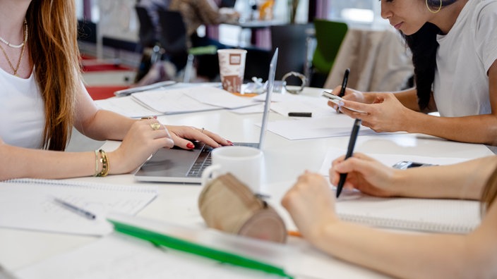 Studierende sitzen um einen Tisch herum, schreiben, tippen in ihre Laptops und schauen auf's Handy. 