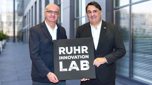 Die Rektoren Manfred Bayer (TU Dortmund) und Martin Paul (RUB) lächeln in die Kamera und halten ein Schild hoch mit der Aufschrift "Ruhr Innovation Lab"