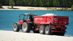 Ein Traktor mit Anhänger fährt eine Ladung Quarzsand auf den Strand nahe der Wasserkante