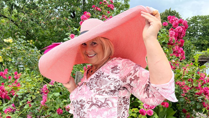 Eine sommerlich-gekleidete Frau mit rosa Hut steht vor einem Rosenstrauch.