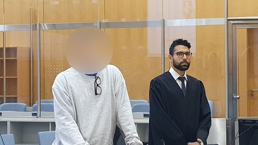 Ein Mann, dessen Gesicht gepixelt ist, steht neben seinem Verteidiger in einem Gerichtssaal.