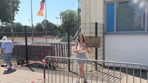 Eine junge Frau steht in einem abgesperrten Wartebereich und hält ein Plakat hoch, auf dem ein spanischer Spruch steht.