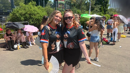 Zwei junge Frauen posieren mit Bad-Bunny-Trikots und Fächern vor einer Warteschlange.
