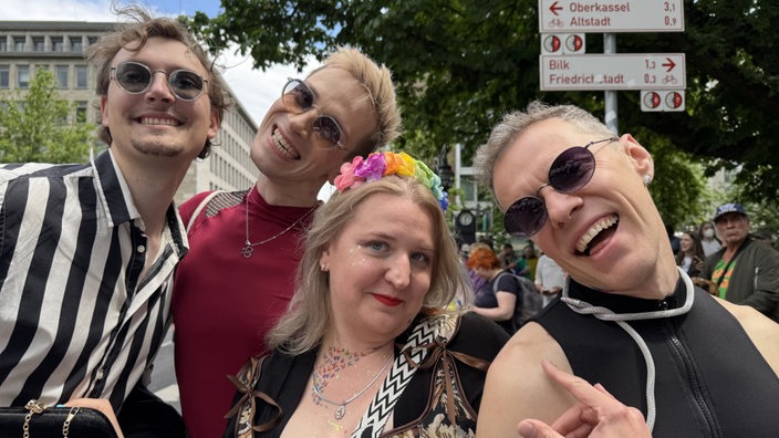 Vier Menschen grinsen in die Kamera beim CSD in Düsseldorf. Die Frau in der Mitte trägt einen Blumenkranz.