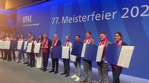 Die Absolventen stehen auf der Bühne in einer Reihe und halten ihre Meisterurkunde in die Kamera