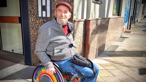 Ein Mann im Rollstuhl schaut in die Kamera. Er sitzt vor einem Haus und die Sonne scheint.