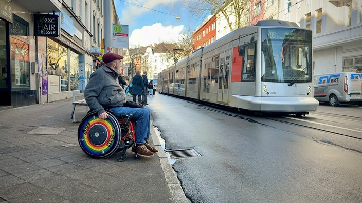 Ein Mann mit Rollstuhl wartet am Bahnsteig auf eine Straßenbahn. Sie fährt gerade ein