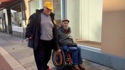 Ein Mann mit Blindenstock und ein Rollstuhlfahrer sind in der Stadt unterwegs.