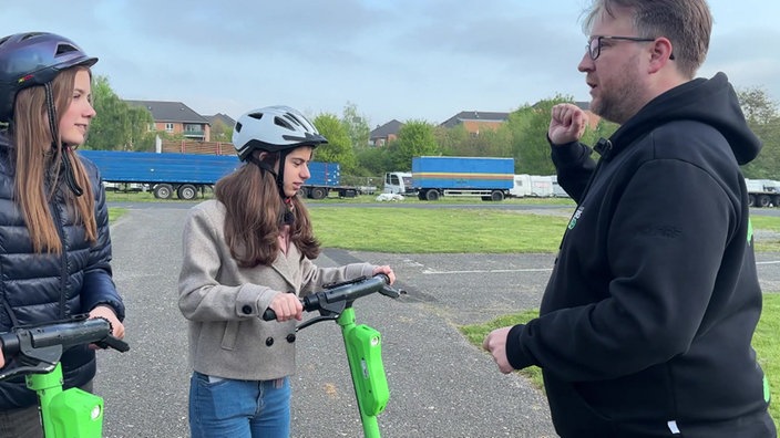 Zwei Schülerinnen stehen auf ihren grünen E-Scootern. Ein Mann im schwarzen Pullover erklärt ihnen wichtige Verkehrsregeln.