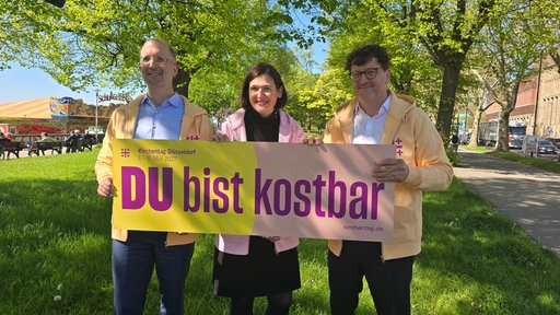 Eine Frau zwischen zwei Männern vor einem lila-gelben Schild mit der Aufschrift "Du bist kostbar"
