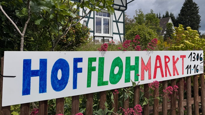 Banner mit der Aufschrift "Hofflohmarkt 13.06 11- 16 Uhr" an einem Zaun befestigt
