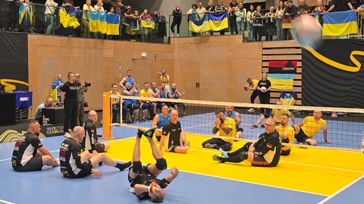 Zwei Mannschaften mit jeweils 6 Personen sitzen durch ein tiefes Volleyball- Netz getrennt. Näher an der Kamera sitzt das schwarz- tragende deutsche Team, weiter hinten die Gelb- tragenden Ukrainer. 