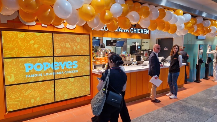Orange weiße Luftballons hängen an der Decke, vorne ein Bildschirm mit der Aufschrift "Popeyes famous louisiana chicken"