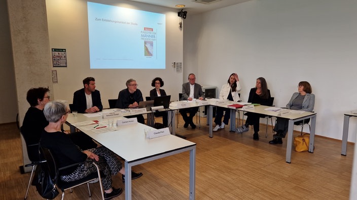 Meherer Personen sitzen an U-förmig zusammengestellten Tischen vor einer Projektionsfläche