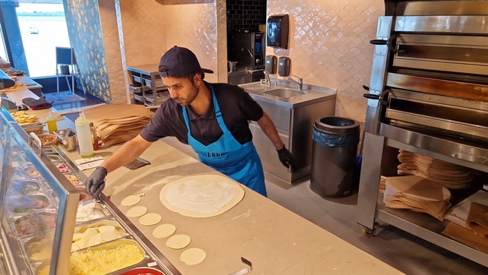 Ein Mann steht hinter einem Tresen und breitet den Teig für eine Pizza aus.