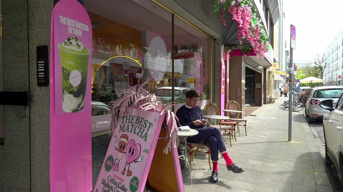 Auf dem Bild sieht man die Außenfassade eines pinken Cafes, davor stehen pinke Stühle, oben hängen pinke Pflanzen. 