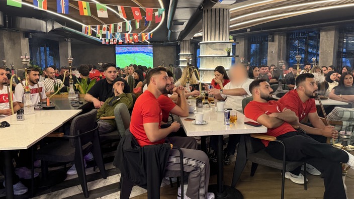 Menschen in Trikots sitzen in einer Gastronomie und gucken auf einen unbestimmten Punkt rechts außerhalb des Bildes