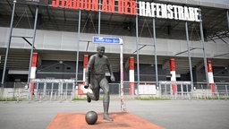 Ein Mann als Bronze-Statue schießt einen Ball. Im Hintergrund ist das RWE Stadion an der Hafenstraße zu sehen