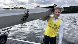Ein Jugendlicher steht vor einem See und trägt ein weißes Kanu auf seiner Schulter