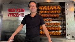 Ümit Yilmaz in schwarzem Poloshirt steht vor einem Grillstand mit mehreren Reihen Brathähnchen im Hintergrund.