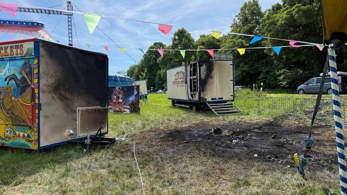 Ein Brandfleck in der Wiese ist umgeben von meheren Zirkuswagen, die zum Teil angebrannt sind.