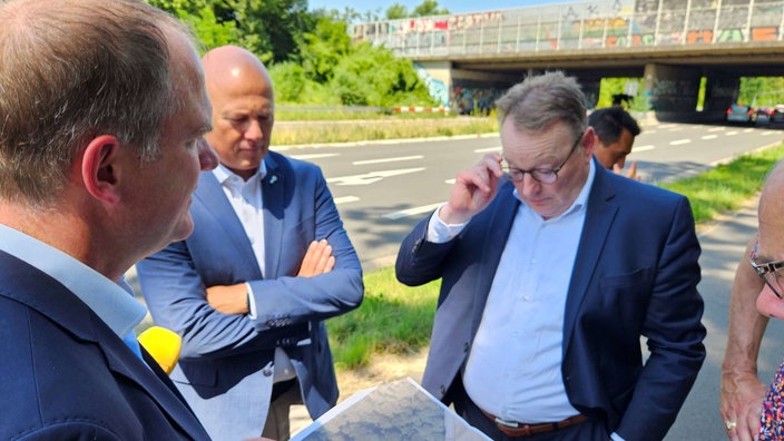 drei Männer (u.A Kölner Oberbürgermeister Torsten Burmester und Hürther Bürgermeister Dirk Breuer) stehen im Kreis neben einer Autobahnbrücke zusammen.