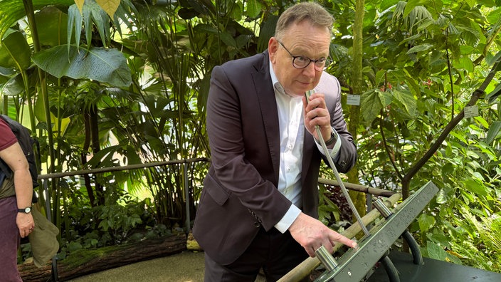 Bürgermeister Burmester erkundet das Gewächshaus und spricht in eine  Art Seil. Dabei drückt er einen Knopf.
