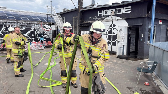 Einsatzkräfte der Feuerwehr transporteiren einen Schlauch. Dahinter das Vereinsheim der wilden Horde (Ultra-Gruppierung 1. Fc. Köln)