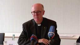Dompropst Guido Assmann sitzt auf einer Pressekonferenz vor einer gestreiften Wand