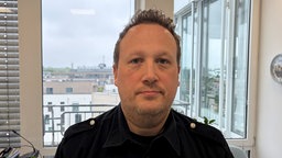 Ein Mann mit kurzen Haaren und dunkler Uniform steht vor dem Fenster in einem Büro und guckt freundlich in die Kamera