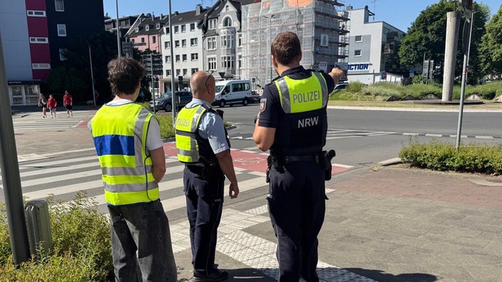 Zwei Polizisten zeigen einem Jungen in Warnweste die Gefahrenstellen am Kreisverkehr.