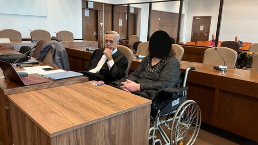 Eine Frau in einem Rollstuhl sitzt neben ihrem Anwalt in einem Gerichtssaal des Kölner Landgerichts