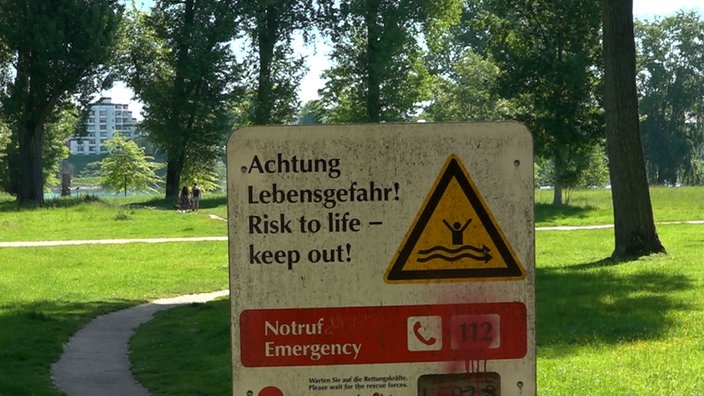 Warnschild an einem Parkweg mit Hinweis auf Lebensgefahr, daneben ein gelbes Dreieck mit einer Person im Wasser.